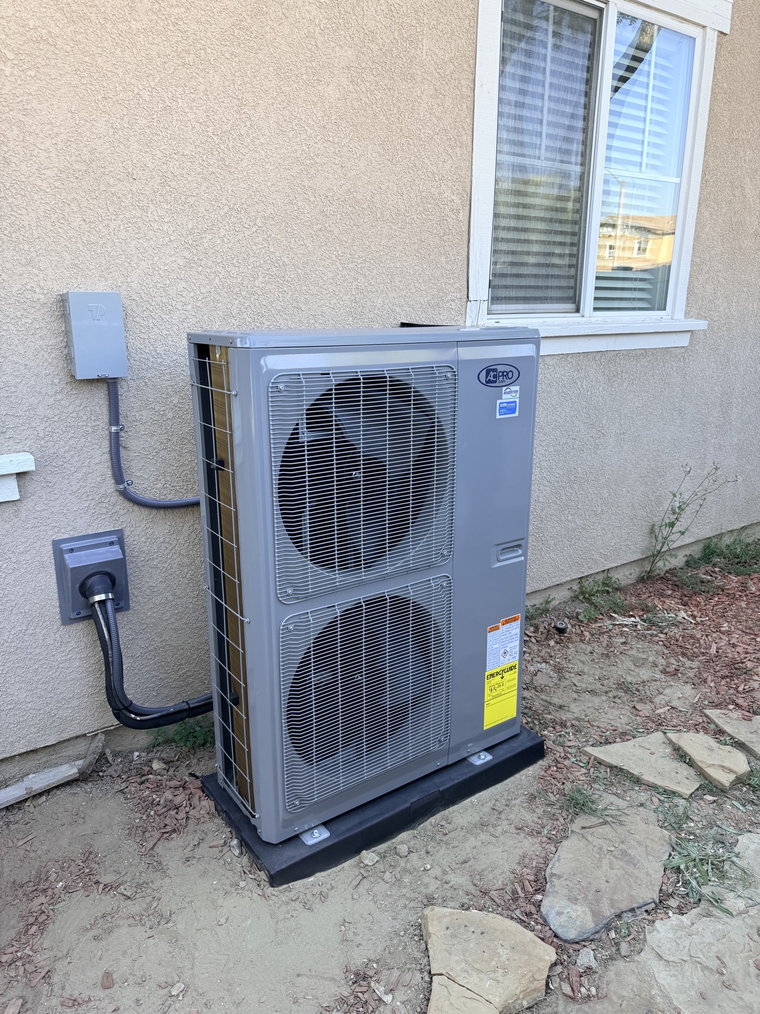 Gray dual-fan condenser on a pad beside a stucco house, disconnect and conduit on the wall